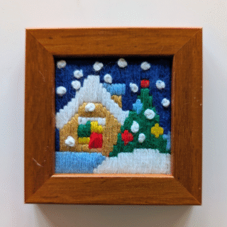Small framed cross-stitch artwork of a snowy cottage with a Christmas tree and falling snow.