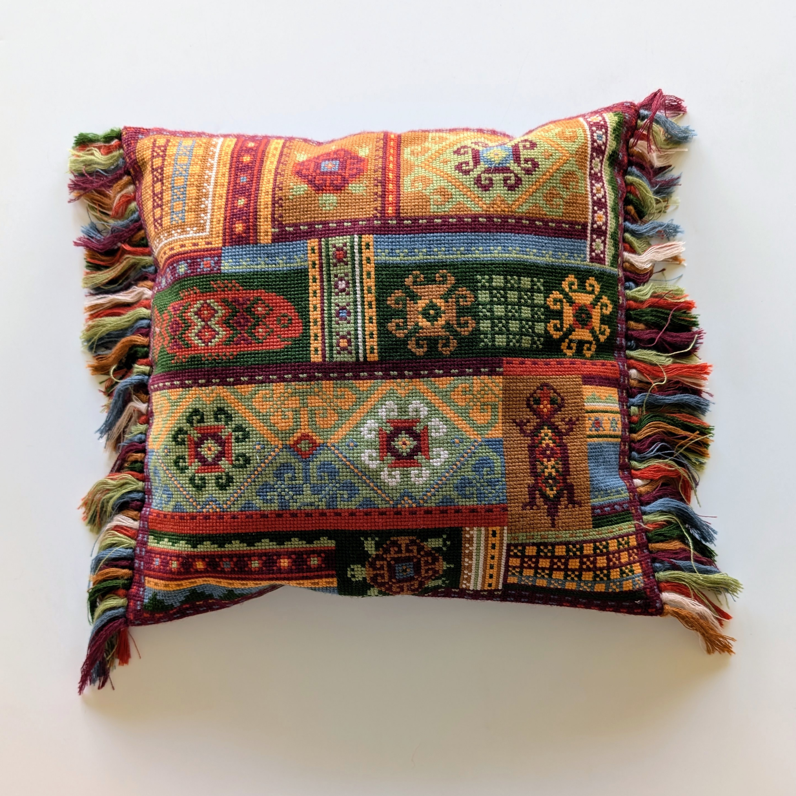 Colorful cross-stitched pillow featuring an oriental-inspired patchwork of geometric motifs, symbols, and bright tassels along the edges.