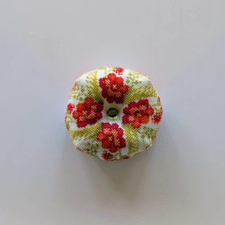 A small round biscornu pincushion stitched with bright red and orange flowers and a decorative bead in the center.