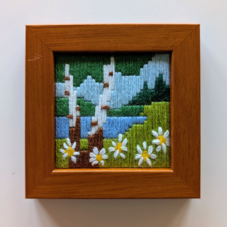 Small framed cross-stitch artwork showing birch trees, a blue river, green hills, and white daisies in the foreground.