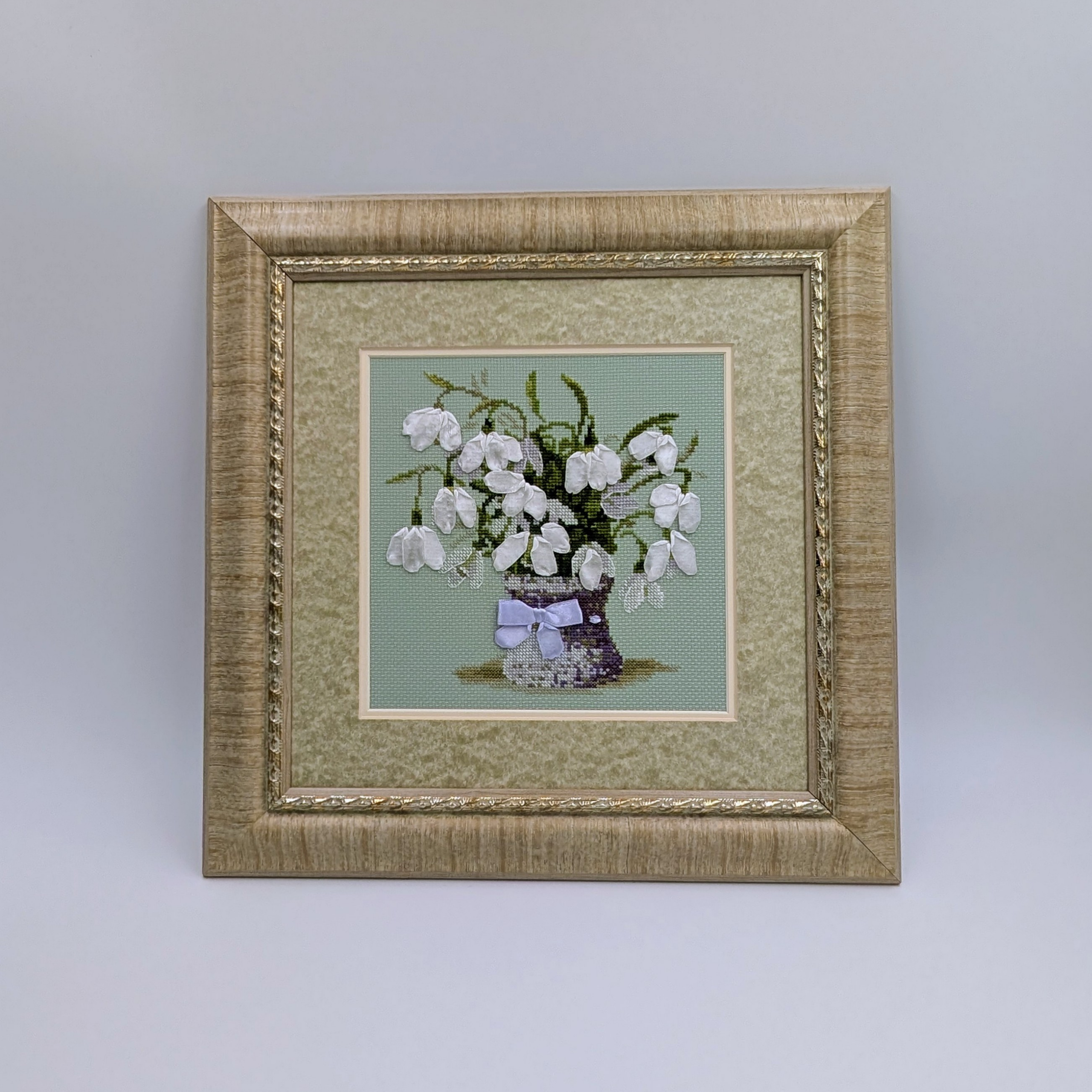 Framed cross-stitch artwork featuring a bouquet of white bellflowers arranged in a purple vase with a delicate ribbon.
