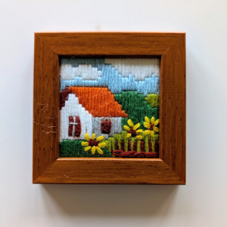 Small framed cross-stitch artwork featuring a white cottage with an orange roof, sunflowers, and a bright countryside background.