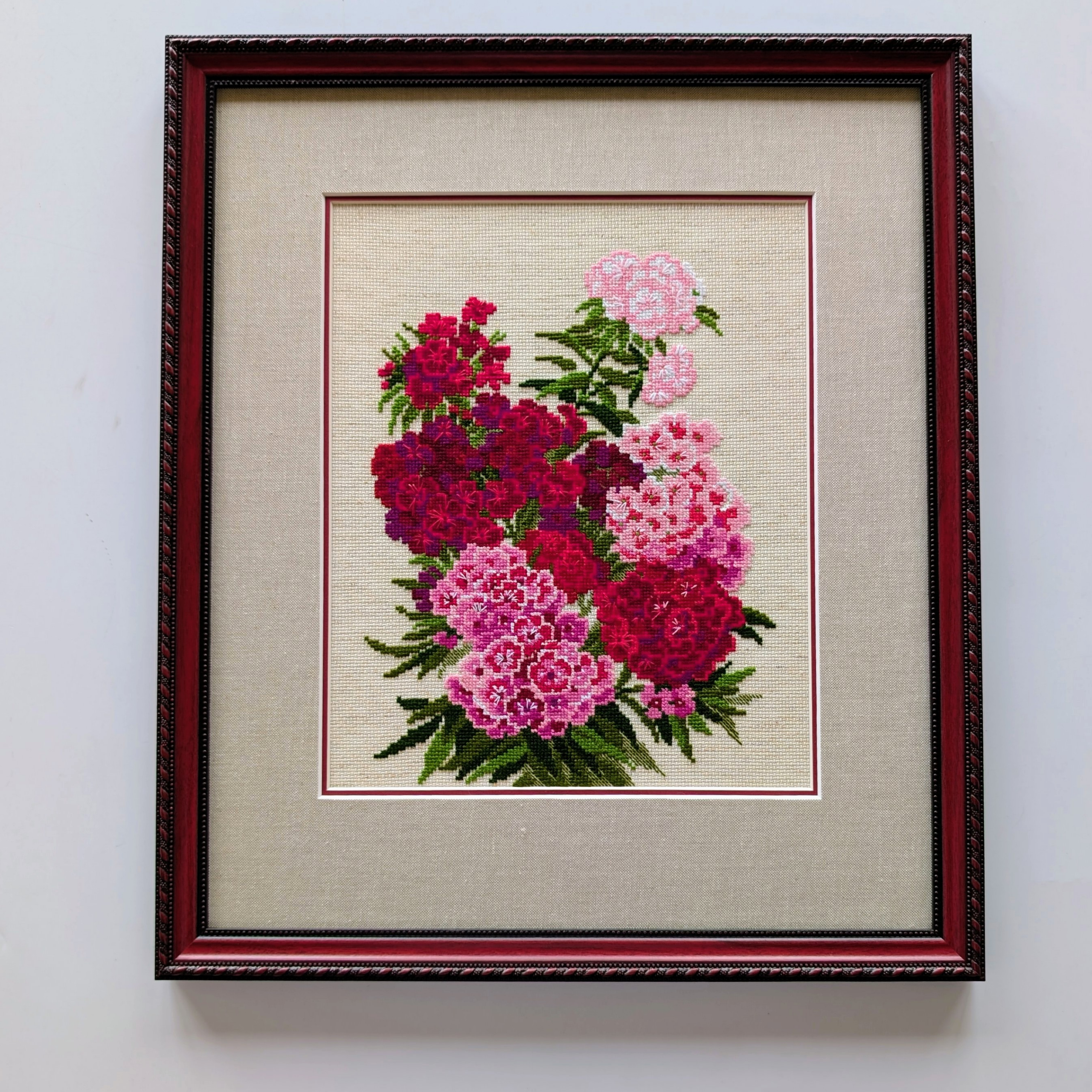 Framed cross-stitch artwork showing a bouquet of bright pink, red, and magenta dianthus flowers with green leaves.