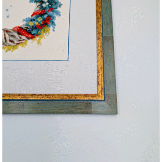 Framed embroidery artwork featuring a colorful winter wreath with pinecones, holly berries, evergreen branches, and a gray-red ribbon.