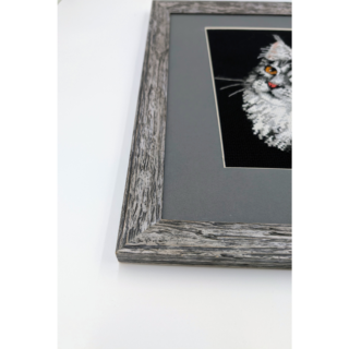 Framed and stitched cross-stitch artwork depicting a Maine Coon cat with expressive amber eyes, fluffy fur, and a curious gaze against a dark background.