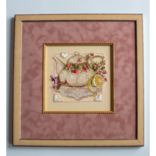 Framed stitched artwork showing a decorative teapot adorned with flowers, lace details, and a lemon on a soft pastel background.